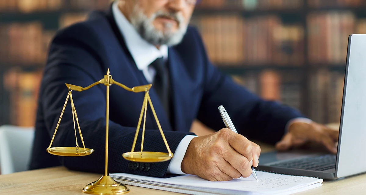 A professional legal expert in a suit taking notes and using a laptop next to a golden scale of justice, representing the Power of Attorney Online Service in Dubai.
