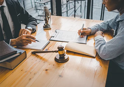 A professional legal advisor sitting at a wooden desk with a client, discussing documents with a gavel and Lady Justice statue in view.
