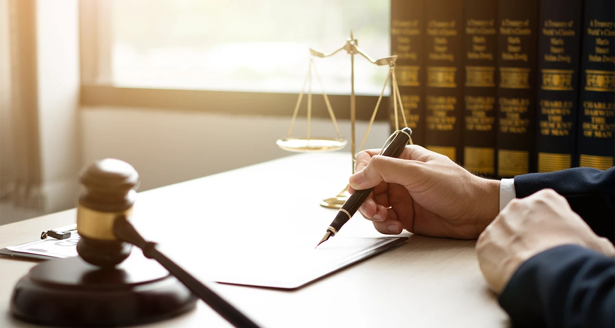 A lawyer's hand signing official legal documents on a desk featuring a judge’s gavel, scales of justice, and law books, representing the process of granting Power of Attorney for Court Case.