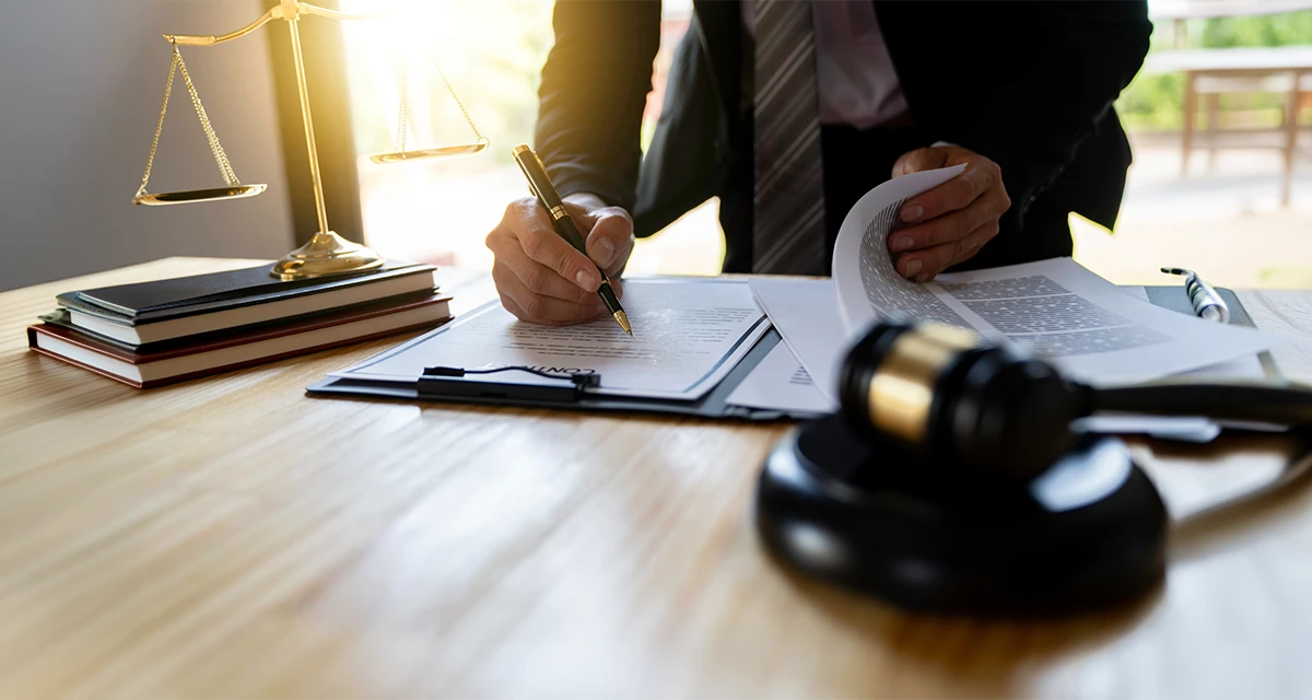 A lawyer in a suit signing legal documents on a wooden desk with a gavel and scales of justice, representing services for an online power of attorney Dubai.